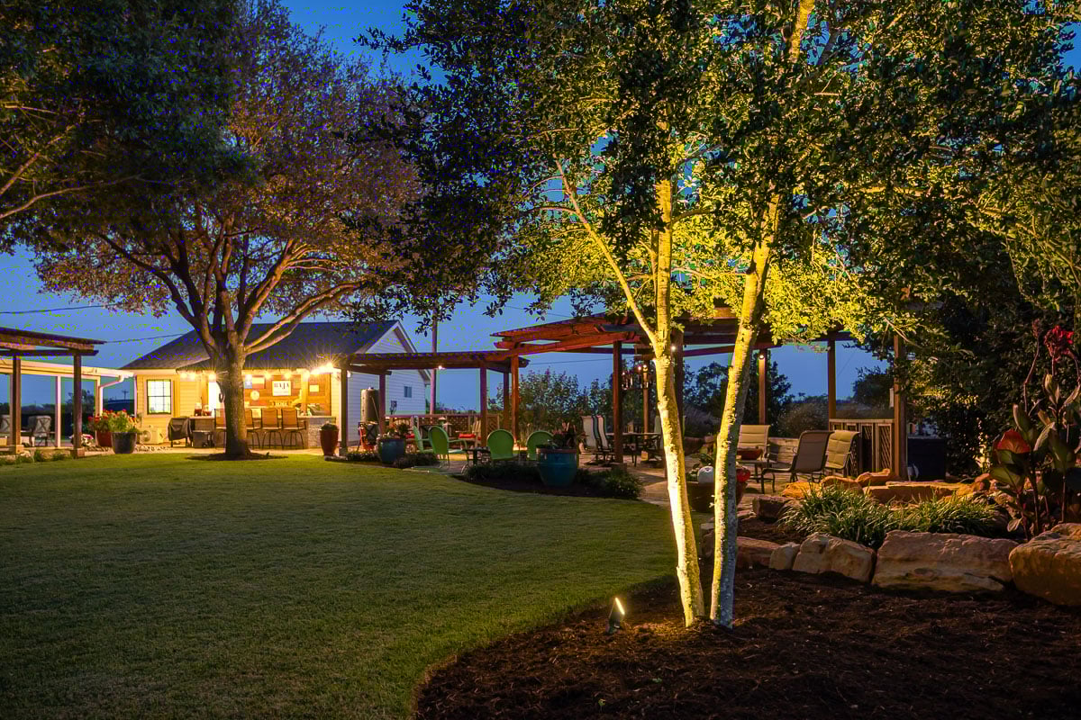 Backyard tree lighting up lighting pergola green lawn outdoor kitchen 